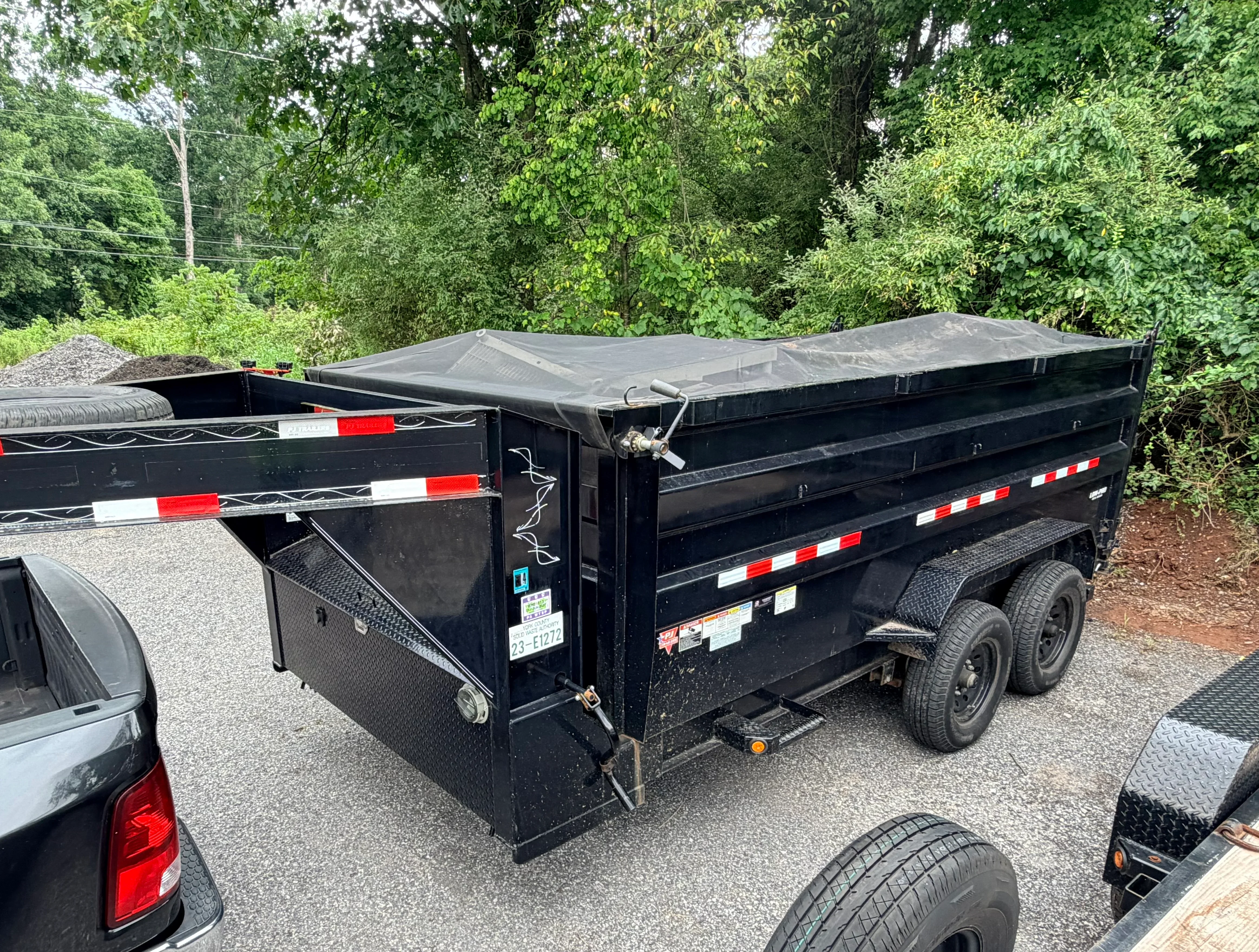 Black dual axle dump trailer with tarp cover and red/white safety stripes